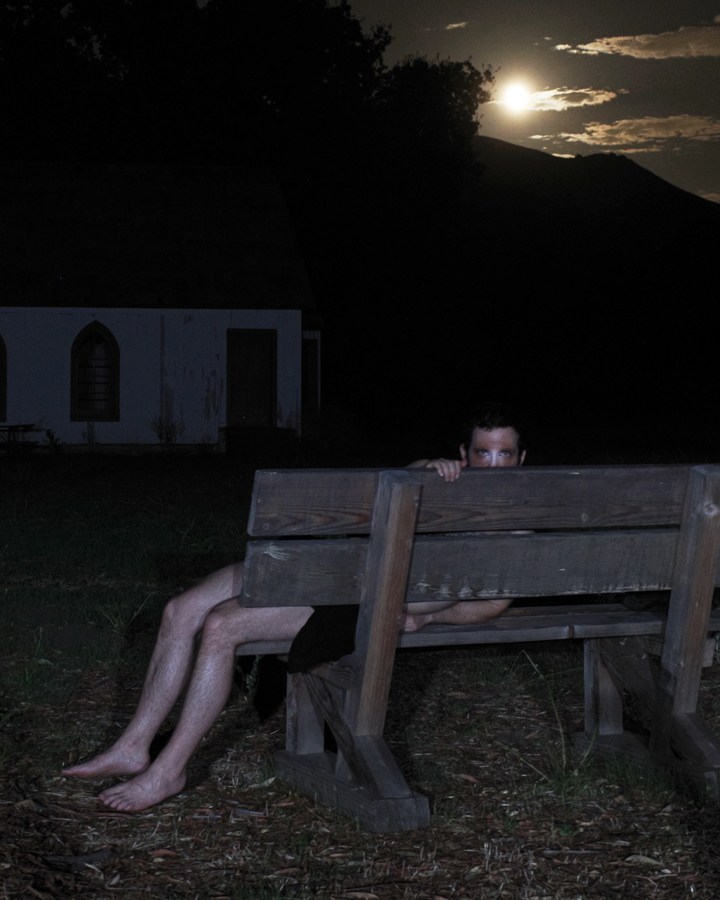 Image of a pale figure reclined on a bench in moonlight with a church behind them peering at the camera.